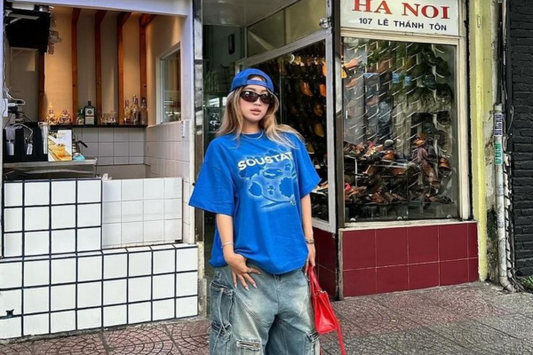 Fashion-forward individual wearing a bold blue graphic oversized T-shirt, distressed baggy jeans with multiple pockets and straps, black sneakers, a matching blue cap worn backwards, and oversized sunglasses. 