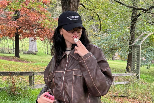 Anna Paul stands outdoors in a park during autumn, wearing a brown leather jacket, blue jeans, and a black cap that reads 'Can I say something without you getting mad.' She is holding a flower to her lips and holding a phone in her other hand. 