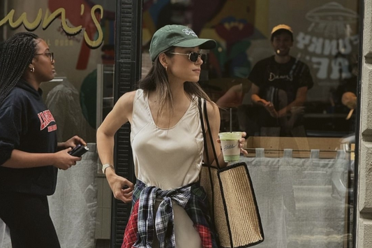 Ruta Zake walks along a New York City sidewalk, wearing a long beige dress with a red and blue plaid shirt tied around her waist. She accessorizes with a green cap, sunglasses, and a large straw tote bag.